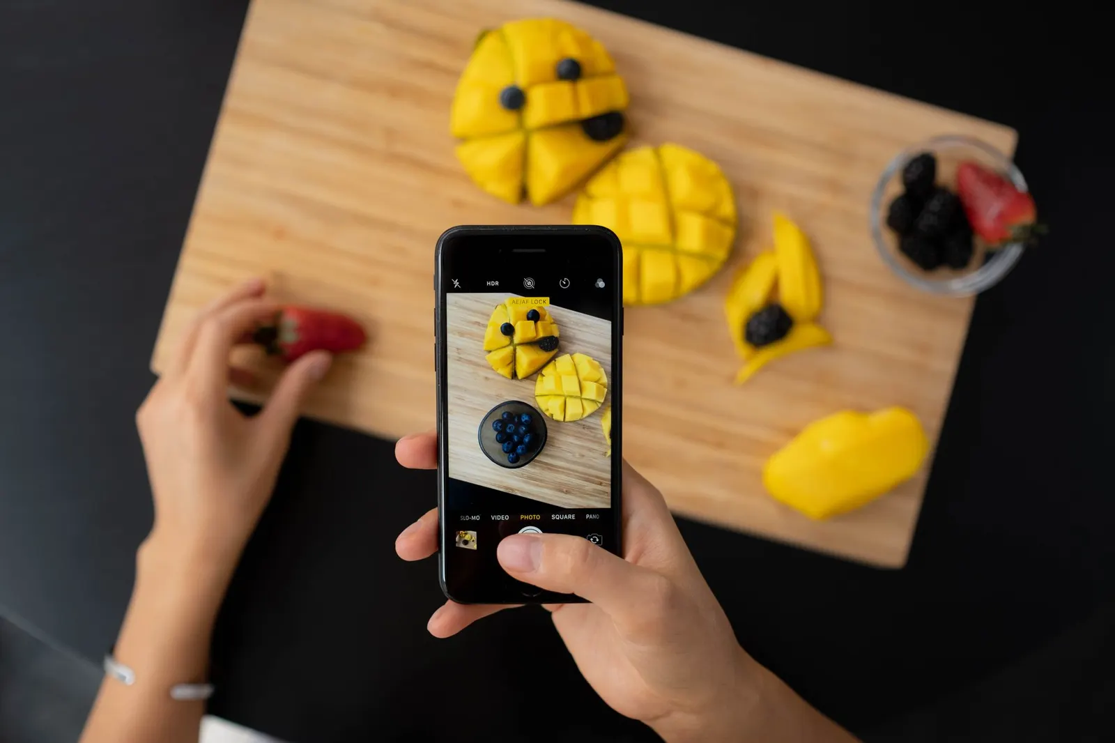 Des mains photographient un plateau de fruits, vu de haut, avec un téléphone.